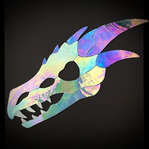 A stained glass iridescent dragon skull cutout. The skull is detailed with teeth and long, pointed horns. The skull is made of a reflective and iridescent glass that creates a rainbow effect.
