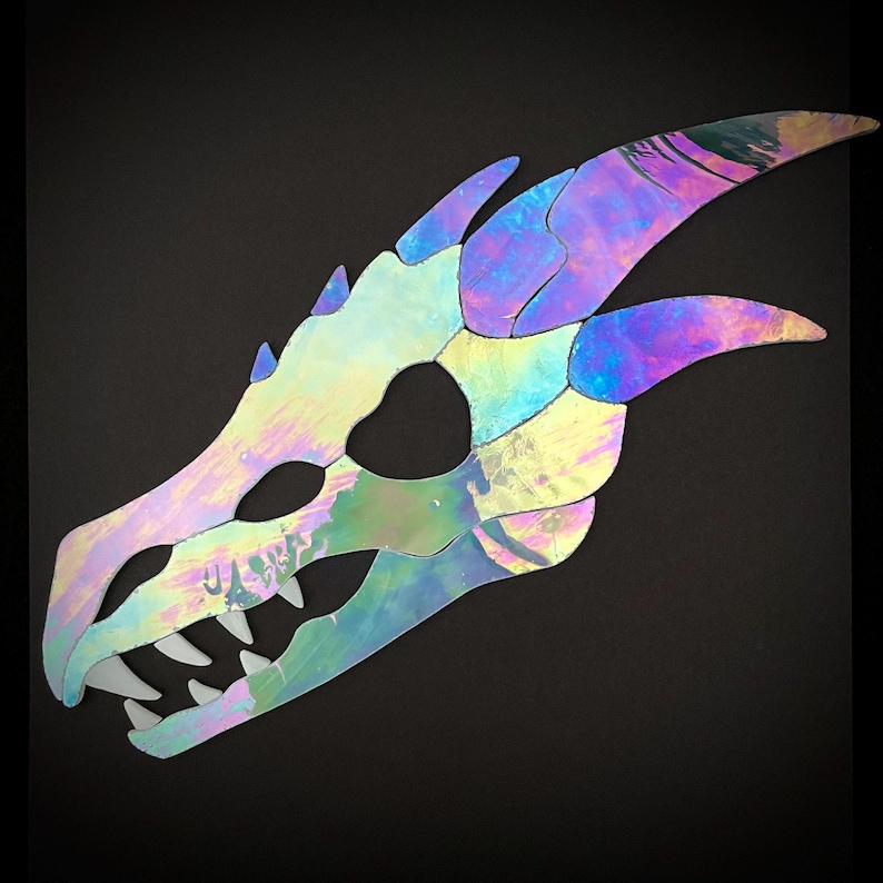 A stained glass iridescent dragon skull cutout. The skull is detailed with teeth and long, pointed horns. The skull is made of a reflective and iridescent glass that creates a rainbow effect.