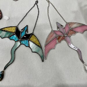 May include: Two stained glass dragonflies hanging from silver chains. One dragonfly is blue, green, and yellow, and the other is pink and white. Both dragonflies have crystal accents.