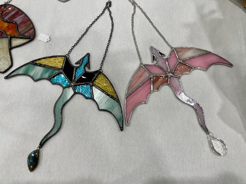 May include: Two stained glass dragonflies hanging from silver chains. One dragonfly is blue, green, and yellow, and the other is pink and white. Both dragonflies have crystal accents.