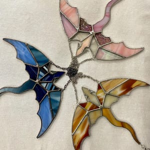 May include: Four stained glass dragonflies hanging from a chain. The dragonflies are in shades of pink, blue, yellow, and brown.