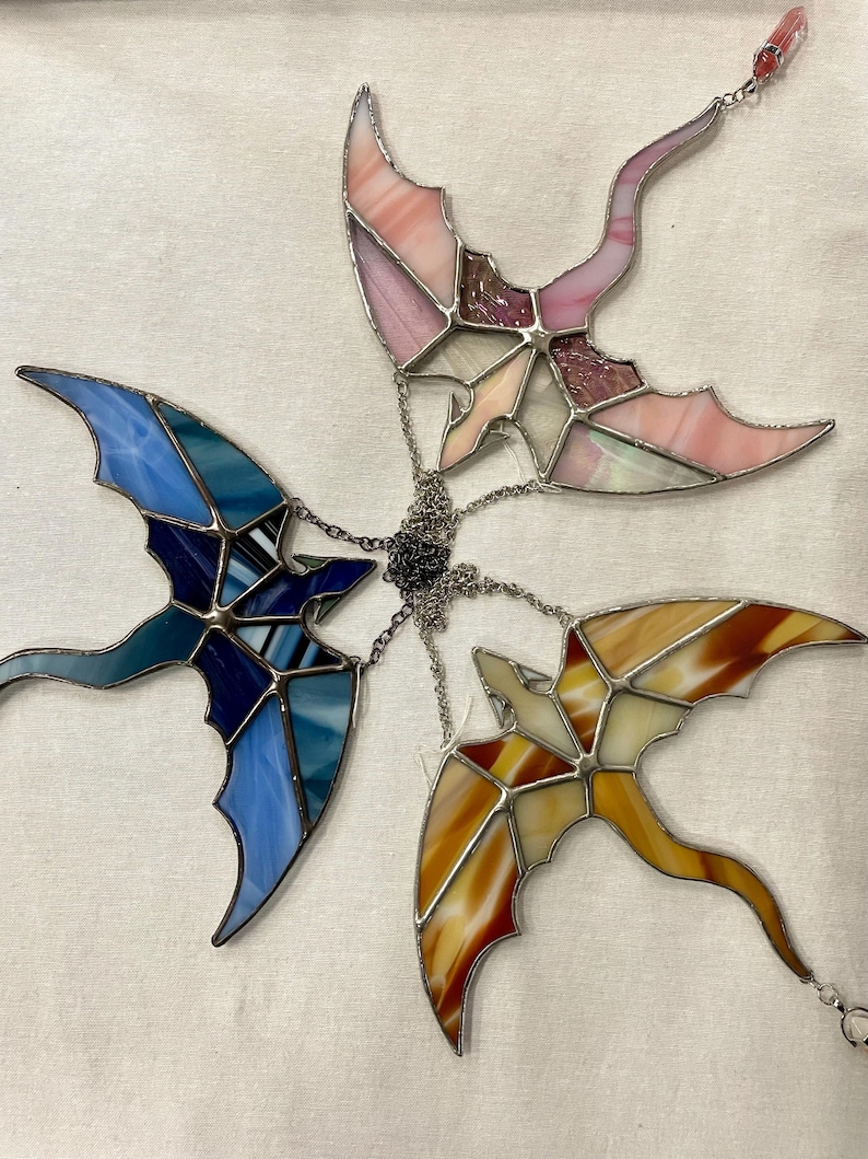 May include: Four stained glass dragonflies hanging from a chain. The dragonflies are in shades of pink, blue, yellow, and brown.