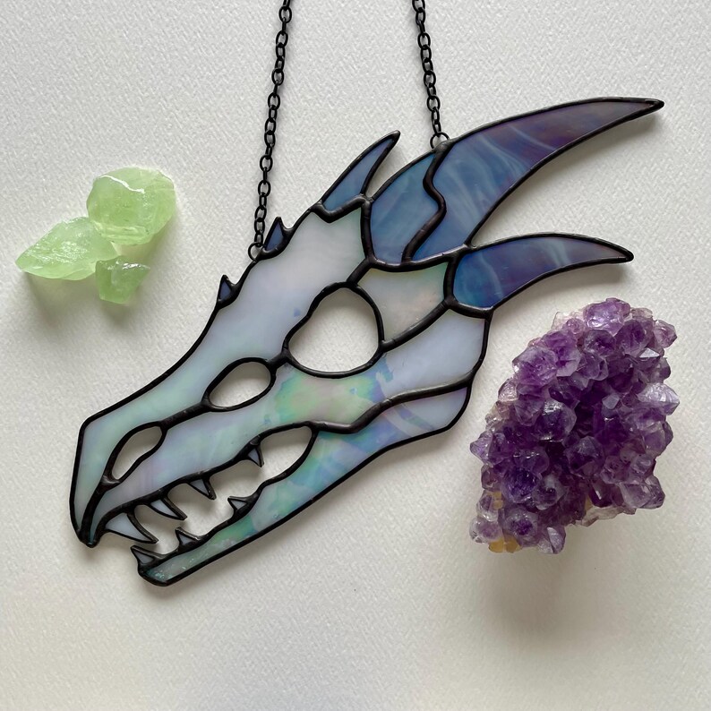 A stained glass dragon skull with iridescent white and grey glass. The skull is hanging from a chain against a white background. The piece is finished in black patina and it is surrounded by green and purple crystals.