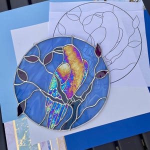 May include: A round stained glass piece featuring a bird perched on a branch, set against a blue background. The bird has iridescent feathers with yellow, purple, and blue hues. The design includes purple leaves and a black outline.