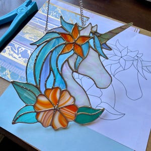 May include: A stained glass unicorn suncatcher featuring iridescent white glass for the head and horn, with a flowing mane of blue and teal glass. An orange and white flower is at the base, and an orange star-shaped flower sits in the mane.