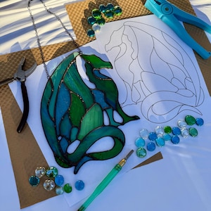 Può includere: Un drago in vetro colorato con sfumature di verde e blu, appeso a una catena, è appoggiato su un tavolo da lavoro. Sono visibili anche strumenti per l'artigianato, perline di vetro e un motivo a drago, che suggeriscono un progetto fai-da-te.