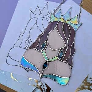 Op de afbeelding: Gebrandschilderd glaskunstwerk dat een koningin met een kroon, lang haar en een hanger afbeeldt. Het stuk heeft tinten paars, blauw en iriserend glas, omlijnd met zilver soldeer. Een ketting is bevestigd om op te hangen. Het kunstwerk staat op een wit oppervlak.