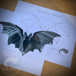 May include: A stained glass bat with marbled black and white patterns placed over a white paper template labelled Flying Bat Pattern. The paper includes the text Rune Glass and a fern-like logo. The bat features segmented glass pieces.