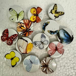 May include: A selection of round, white buttons, each decorated with a different butterfly design. The butterflies are depicted in a variety of colours, including yellow, orange, red, blue, and black. The buttons are arranged on a white surface.