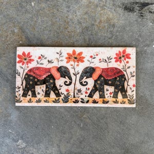 May include: A rectangular ceramic tile with a white background and a floral design featuring two black elephants with red and orange accents. The elephants are facing each other and have their trunks raised.