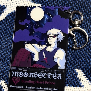 Peut inclure: Un porte-clés rectangulaire en acrylique représentant un personnage aux cheveux blancs et lunettes de soleil sombres sous un ciel nocturne avec une pleine lune. Le texte indique MOONSETTER, Bleeding Heart Prince et Show ticket :: Land of tombs and krypton.