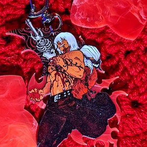 May include: A clear acrylic keyring featuring a stylised illustration of a shirtless character with white hair, dark trousers, and a skeletal figure behind them. The character has red markings on their torso and is holding a red heart-shaped object.