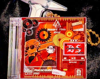 May include: A red and orange Mini Disc MD case with a cartoon graphic, a silver charm, and a gold chain. The case has the text "Insert This End" and "Doccond". The image also includes small decorative pieces.