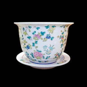 May include: A white ceramic planter with a flared rim and a matching saucer, decorated with a colourful floral pattern featuring pink peonies, green leaves, and small blue and yellow blossoms scattered across the exterior surface of the pot.
