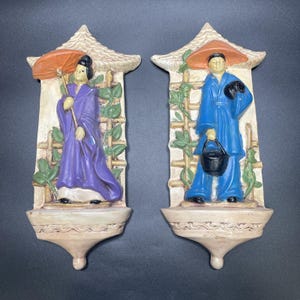 May include: Pair of Asian-themed wall plaques. One features a figure in a purple robe holding an orange umbrella. The other depicts a figure in a blue robe with a black hat and bucket. Both are set against a trellis with green leaves.