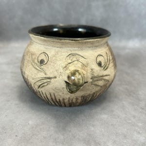 May include: A small, round ceramic bowl with a whimsical face design. The bowl is beige with a dark brown interior and features etched details for eyes, whiskers, and a nose. The overall design gives the bowl a playful, animal-like appearance.