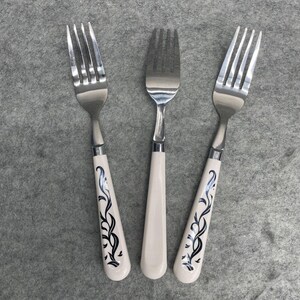 May include: Three stainless steel forks with off-white handles. Two forks have a black, stylized floral design on the handle. The third fork has a plain off-white handle. The forks are arranged on a gray surface.
