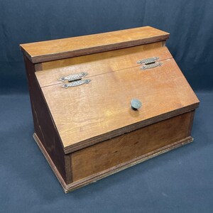 Può includere: Una scatola di legno con coperchio inclinato e un piccolo pomello. La scatola è realizzata in legno marrone chiaro con accenti marrone scuro. Ha due cerniere decorative in metallo e un piccolo pomello per l'apertura.