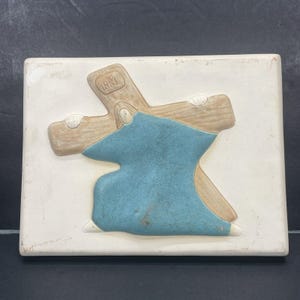 Op de afbeelding: Een rechthoekige, crèmekleurige keramische tegel met een reliëfsculptuur van een figuur die een houten kruis draagt. Het kruis is lichtbruin met de letters INRI. De figuur draagt een blauwe jurk. De tegel heeft een verweerde uitstraling.