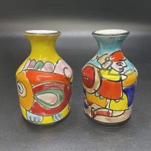 May include: Two hand-painted ceramic vases. One vase is yellow with a fish design in red, orange, and green. The other vase is blue with a figure design in red, yellow, and blue. Both vases have a black rim.