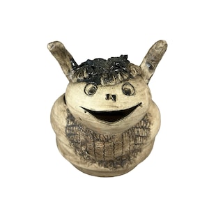 May include: A whimsical ceramic creature with a smiling face, large eyes, and two upright ears. The sculpture has a textured, off-white body with dark accents on the hair and details. The creature's open mouth reveals a hollow interior.