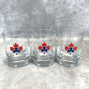 Puede incluir: Tres vasos transparentes con un diseño de hoja de arce roja y trébol azul. El texto "Canadian Club" está impreso debajo. Los vasos son cilíndricos y se apoyan sobre una superficie gris claro.