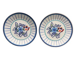 May include: Two white ceramic plates with a blue and white striped border. Each plate features a whimsical fish design with a blue flower, red accents, and a light blue and white striped pattern.