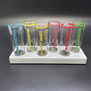 May include: A set of six clear glass shot glasses arranged in a white rectangular holder. Each glass features a unique cocktail name printed in a specific color, including Manhattan, Screwdriver, Bloody Mary, Tom Collins, Margarita, and Black Russian.