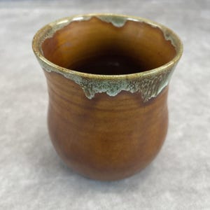May include: A handmade ceramic mug with a rounded base and a slightly flared rim. The mug is a warm brown color with a textured surface. The rim has a unique, dripped glaze in shades of green and brown.
