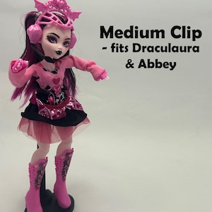 May include: A pink doll with black and pink hair, wearing a pink outfit with a black skirt and pink boots. The doll has a pink headpiece and headphones. Text on the image reads "Medium Clip - fits Draculaura & Abbey."