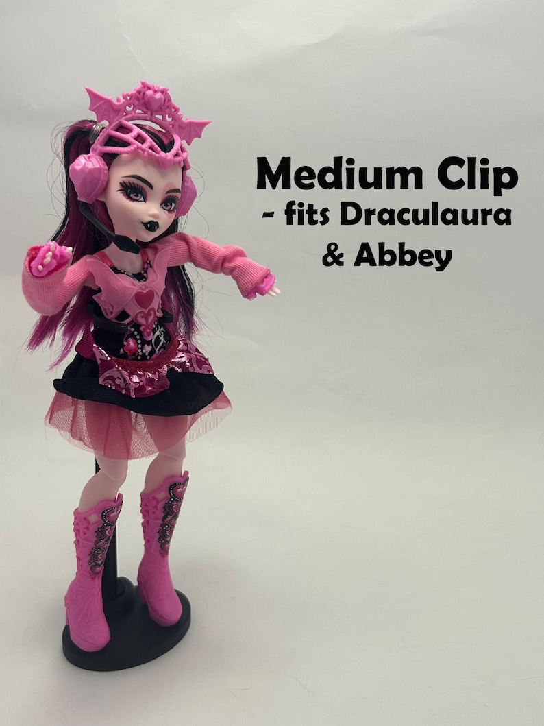 May include: A pink doll with black and pink hair, wearing a pink outfit with a black skirt and pink boots. The doll has a pink headpiece and headphones. Text on the image reads "Medium Clip - fits Draculaura & Abbey."