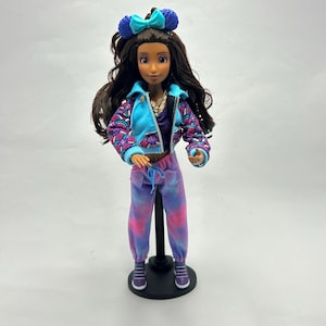 May include: A doll with long brown hair, wearing a blue headband with a bow, a blue jacket with a patterned design, a purple top, and tie-dye pants. The doll is standing on a black stand.