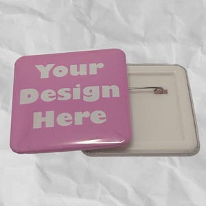 May include: A square pink button with the text "Your Design Here" in white. The button is partially detached, revealing the pin backing. The background is a crumpled white surface.