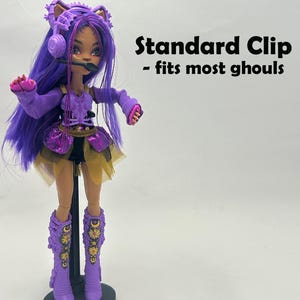 May include: A doll with purple hair, wearing a purple outfit with gold accents, and matching boots. The doll has a headset microphone and is standing on a black stand. Text on the right reads "Standard Clip - fits most ghouls."