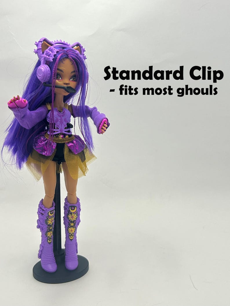 May include: A doll with purple hair, wearing a purple outfit with gold accents, and matching boots. The doll has a headset microphone and is standing on a black stand. Text on the right reads "Standard Clip - fits most ghouls."