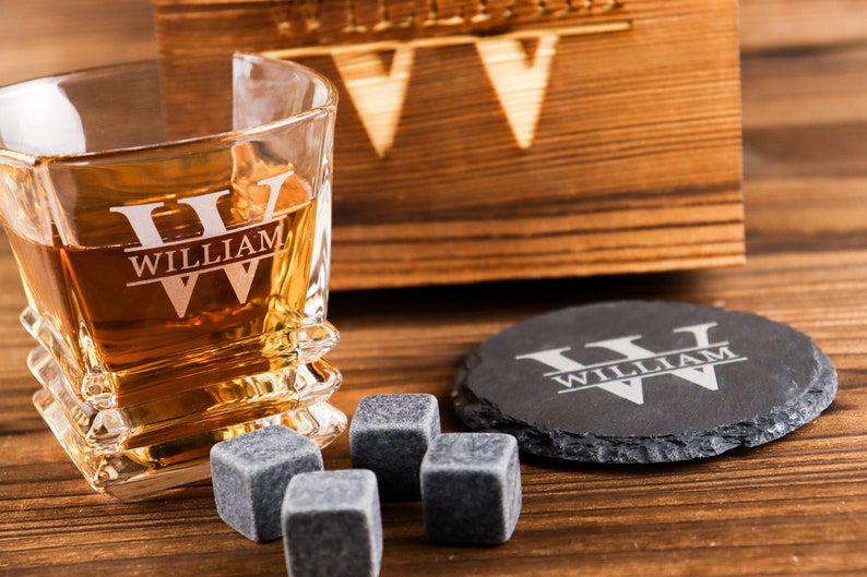Puede incluir: Un juego de vasos de whisky personalizado con una caja de madera, un posavasos de pizarra y cuatro piedras de whisky. El vaso y el posavasos est&aacute;n grabados con el nombre "William" y las iniciales "W" en un estilo de monograma dividido.
