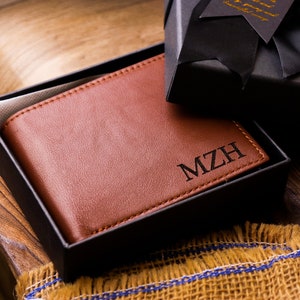 May include: A brown leather wallet with the initials "MZH" engraved on the front. The wallet is in a black gift box.