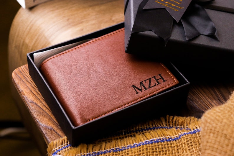 May include: A brown leather wallet with the initials "MZH" engraved on the front. The wallet is in a black gift box.