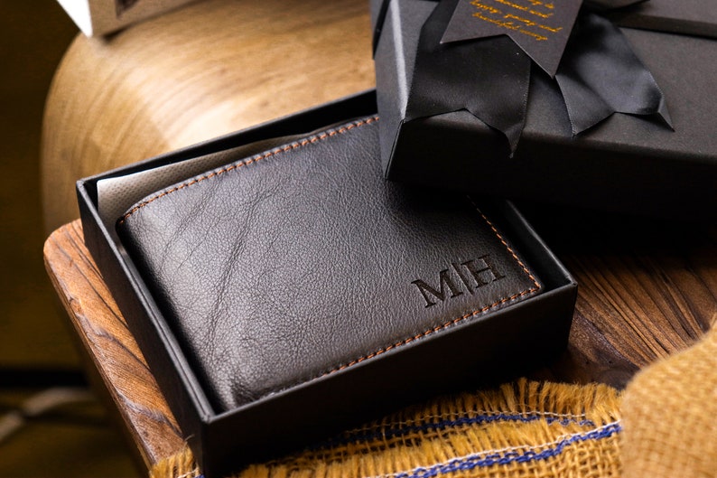 May include: A black leather wallet with orange stitching in a black gift box. The wallet has the initials "MH" embossed on the front.