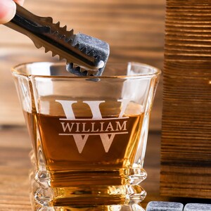 Puede incluir: Un vaso de whisky personalizado con el nombre "William" grabado en &eacute;l. El vaso est&aacute; lleno de l&iacute;quido &aacute;mbar y se coloca una piedra de whisky en el vaso con unas pinzas.