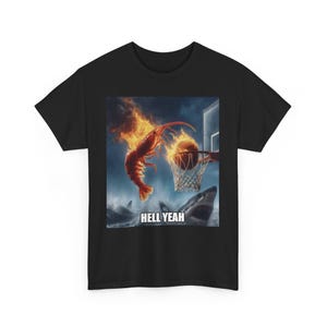 以下が含まれることがあります： 黒いTシャツ。燃えるエビがバスケットボールをダンクするユーモラスなグラフィックが特徴で、背景にはサメがいます。「HELL YEAH」というテキストが画像の下に表示されています。