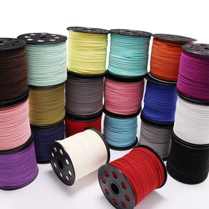 Op de afbeelding: Assortiment van spoelen met gekleurde suède koord, elk gewikkeld rond een zwarte plastic spoel met een metalen wikkelmechanisme. De koorden zijn in verschillende kleuren, waaronder rood, blauw, groen, paars, bruin, roze, wit en zwart.