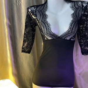 May include: Black lingerie set on a mannequin. The top features a black bodice with a deep V-neck and lace sleeves. A matching thong is placed in front of the mannequin. The background is a neutral-toned fabric.