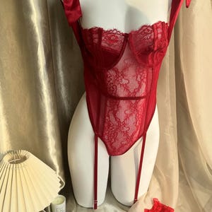 May include: A red lingerie set on a mannequin. The set includes a lace-detailed bodysuit with garter straps and a pair of matching thigh-high stockings. The bodysuit has satin ribbon shoulder straps tied into bows.