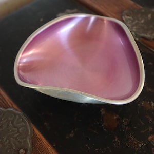 Puede incluir: Un pequeño cuenco de metal de forma triangular con un acabado interior rosa metálico liso y un borde exterior de tono plateado. El cuenco presenta bordes orgánicos suavemente curvados y una base poco profunda, mostrando una superficie pulida y reflectante.