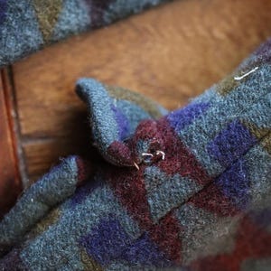 Może przedstawiać: Odzież z grubego, filcowanego materiału wełnianego z geometrycznym wzorem tr&oacute;jkąt&oacute;w w odcieniach ciemnej szarości, głębokiego fioletu, oliwkowej zieleni i bordo. Materiał ma teksturowaną, puszystą powierzchnię z widocznymi szwami na krawędziach.