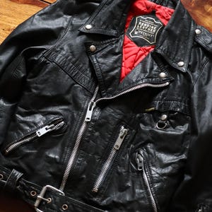 Könnte beinhalten: Eine schwarze Motorradjacke aus Leder mit leuchtend rotem Steppfutter, silberfarbenen Metallreißverschlüssen und einem Taillengürtel mit Metallschnalle. Das Innenetikett lautet: This jacket is manufactured from GENUINE LEATHER for your total comfort PETROFF.