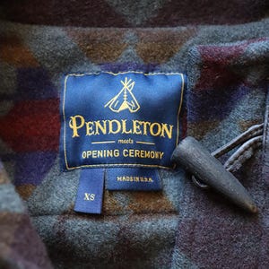 Może przedstawiać: Wełniany płaszcz w geometryczny wz&oacute;r w odcieniach szarości, fioletu i brązu. Niebieska metka zawiera napis Pendleton meets OPENING CEREMONY, MADE IN U.S.A. oraz XS. Ciemne drewniane zapięcie typu kołek jest przymocowane sk&oacute;rzaną pętelką.