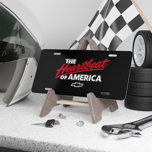 Puede incluir: Placa de matrícula negra con el texto "THE Heartbeat OF AMERICA" en rojo y blanco, y un logotipo de Chevrolet. La placa se muestra sobre un soporte de madera, con una bandera de carreras, neumáticos y herramientas en el fondo.