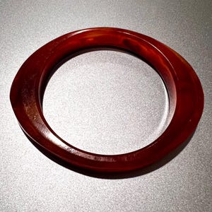 Op de afbeelding: Een gepolijste, roodbruine armband. De armband heeft een glad, afgerond ontwerp met een licht ovale vorm. De kleur varieert van een diep, donkerrood tot lichtere tinten, waardoor een gemarmerd effect ontstaat. Een eenvoudige, elegante accessoire.